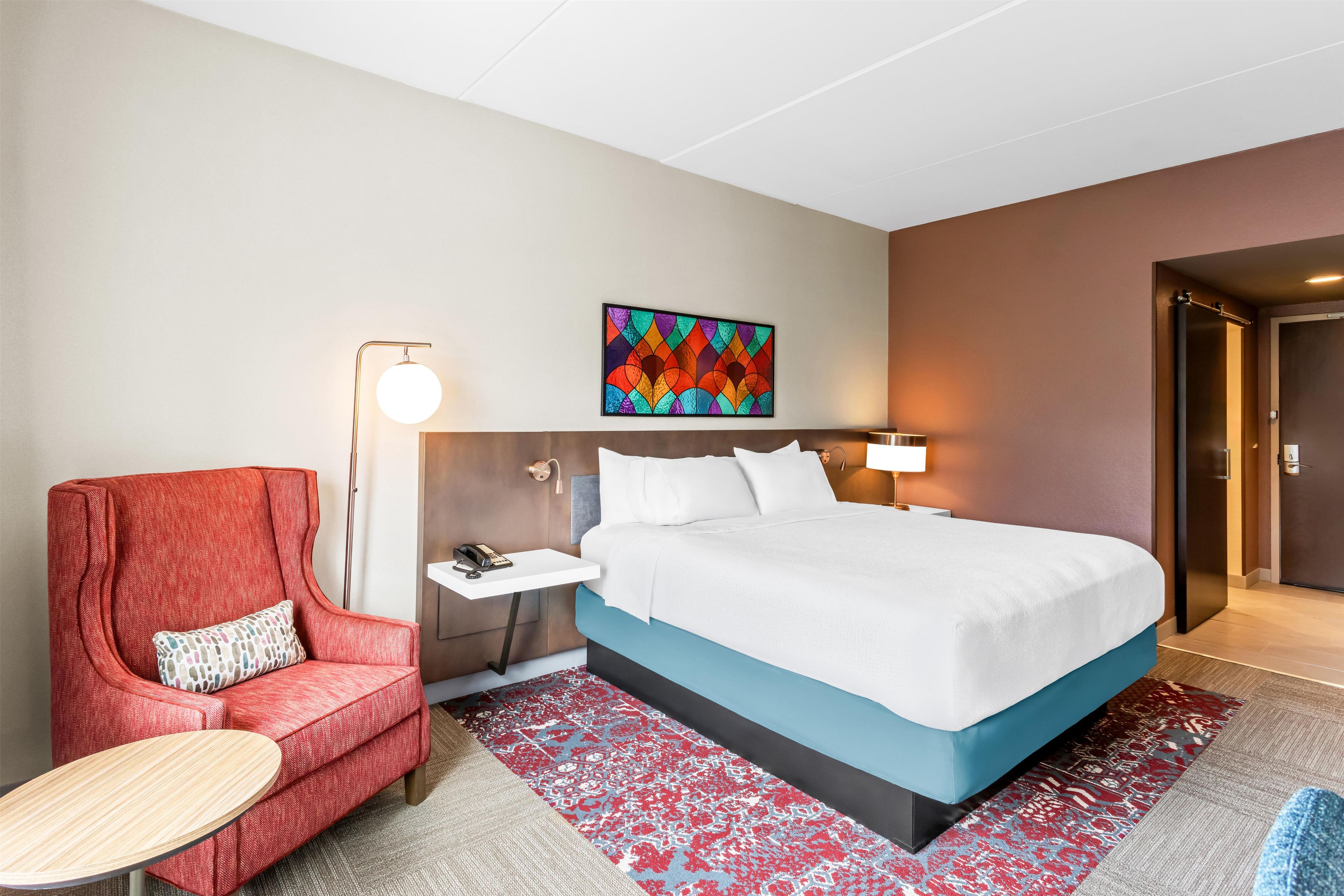 Ein Hotelzimmer mit einem Bett, einem roten Sessel, einem kleinen Tisch, einem Nachttisch, einer Wandleuchte und einem farbenfrohen Kunstwerk über dem Bett.
