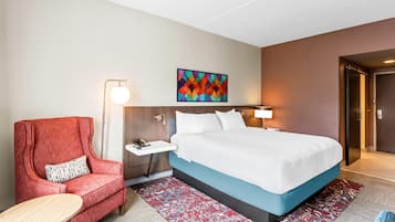 Ein Hotelzimmer mit einem Bett, einem roten Sessel, einem kleinen Tisch, einem Nachttisch, einer Wandleuchte und einem farbenfrohen Kunstwerk über dem Bett.