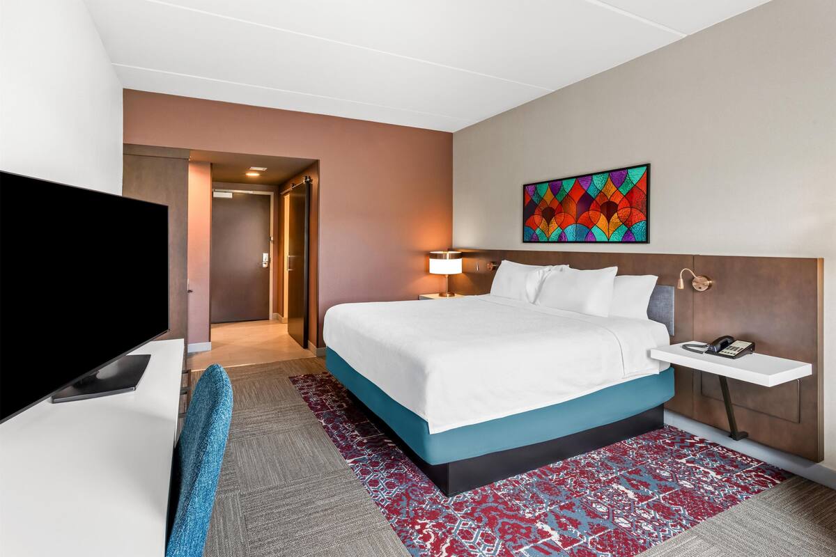 Ein Hotelzimmer mit einem Bett, einem Fernseher, einem Schreibtisch, einer Lampe und einem farbenfrohen Kunstwerk an der Wand.