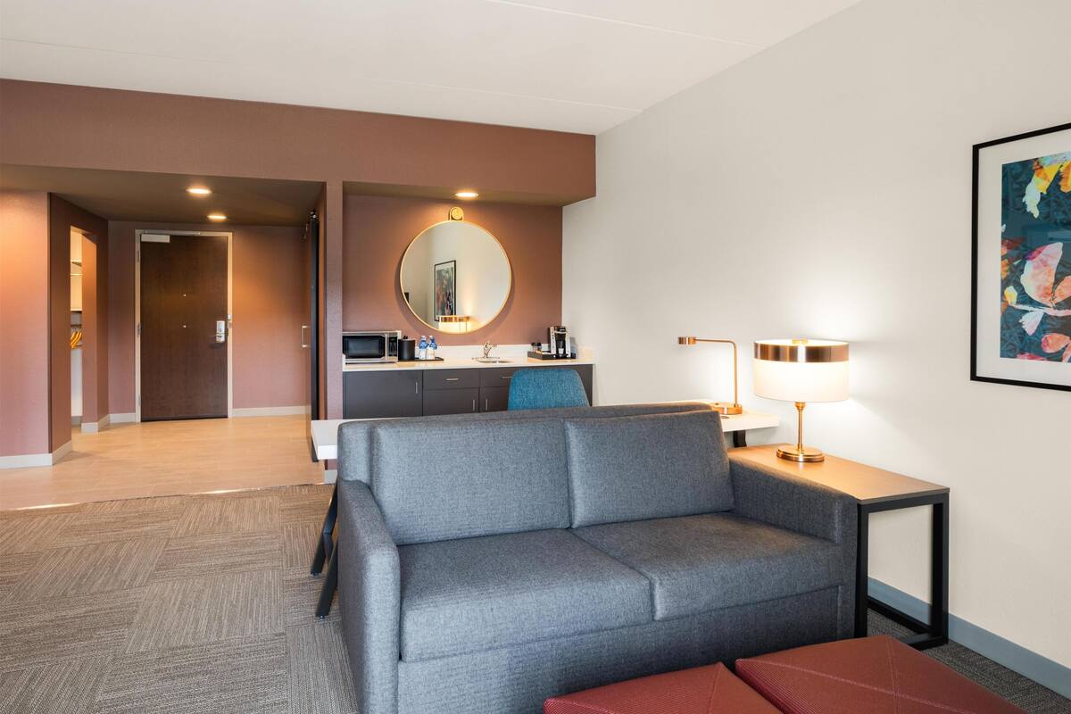 Ein modernes Hotelzimmer mit grauem Sofa, einem kleinen Tisch mit Lampe, einem runden Spiegel und einem Gemälde an der Wand.