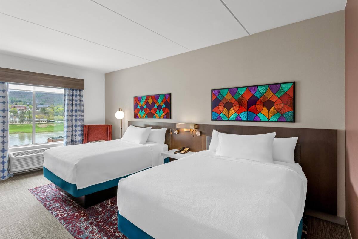 Ein Hotelzimmer mit zwei Betten, einem großen Fenster mit Blick und zwei farbenfrohen Kunstwerken an der Wand.