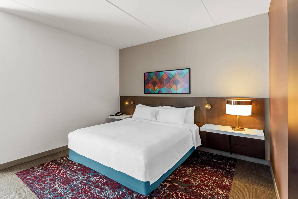 Ein Hotelzimmer mit Bett, Nachttisch, Lampe, Wandbild und einem gemusterten Teppich.