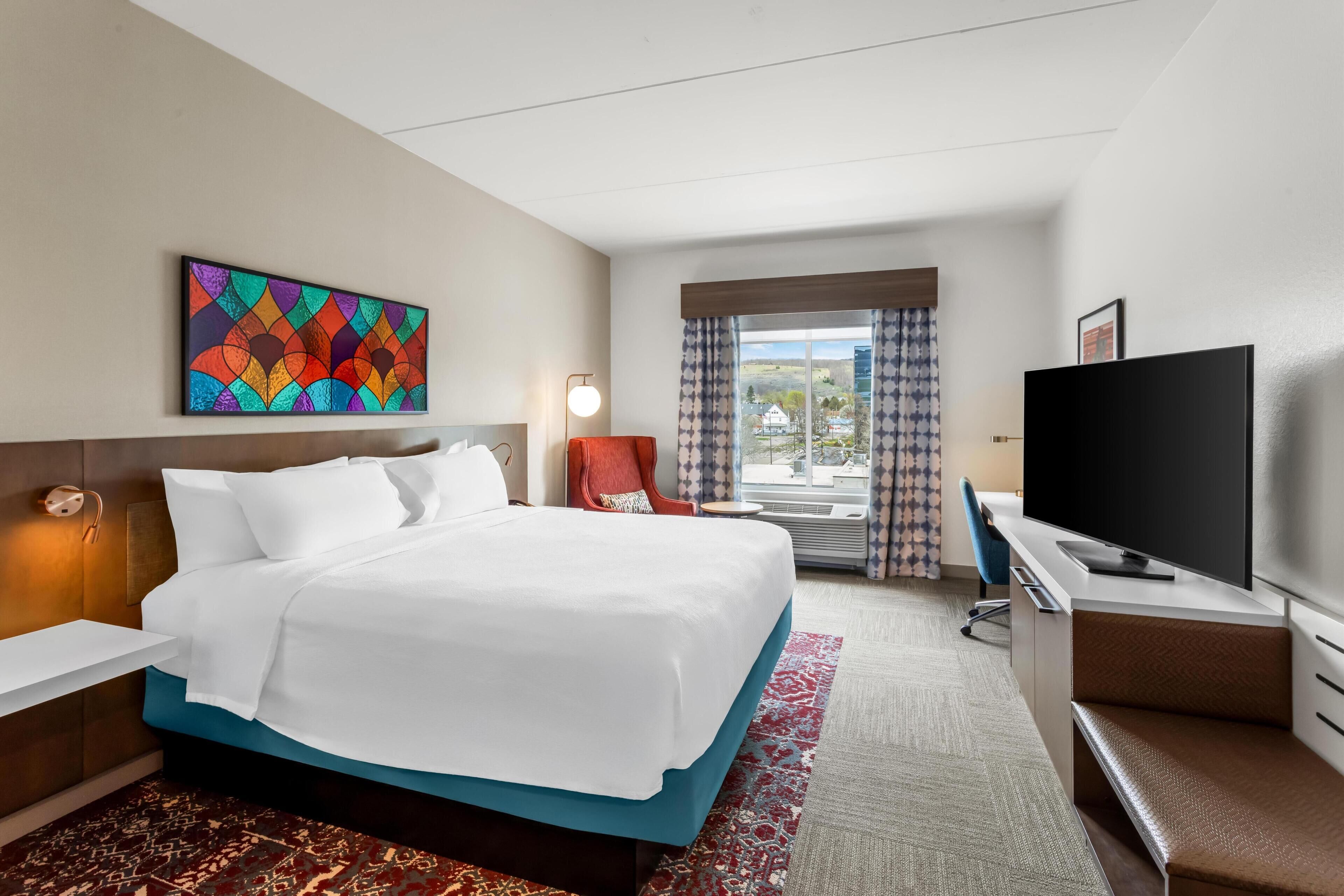 A hotel room with a bed, a desk, a television, a painting on the wall, and a window with curtains.