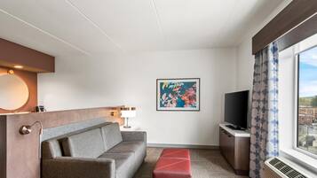 A hotel room with a sofa, a flat-screen TV, a painting, and a window with curtains.