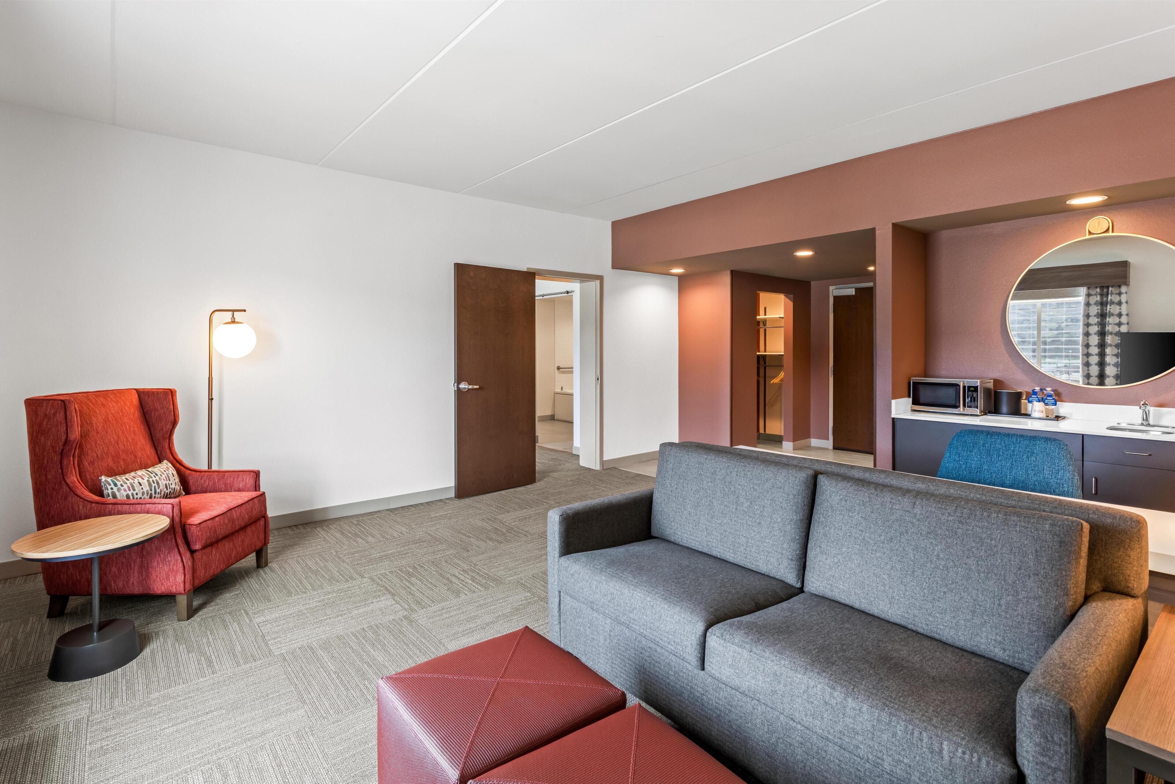 A modern hotel room with a grey sofa, red armchair, and a small round table.