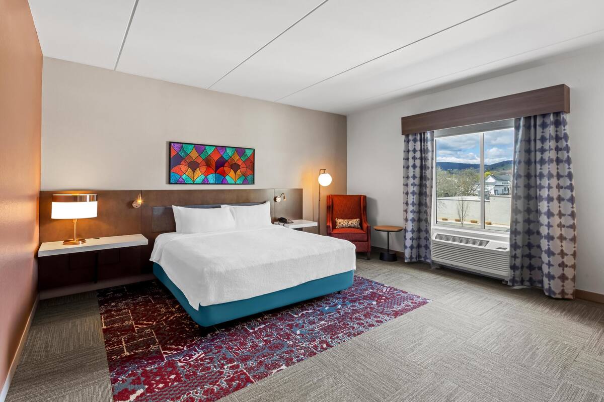 Ein Hotelzimmer mit Bett, Nachttisch, Lampe, Stuhl, Fenster mit Vorhängen und einem farbenfrohen Kunstwerk an der Wand.