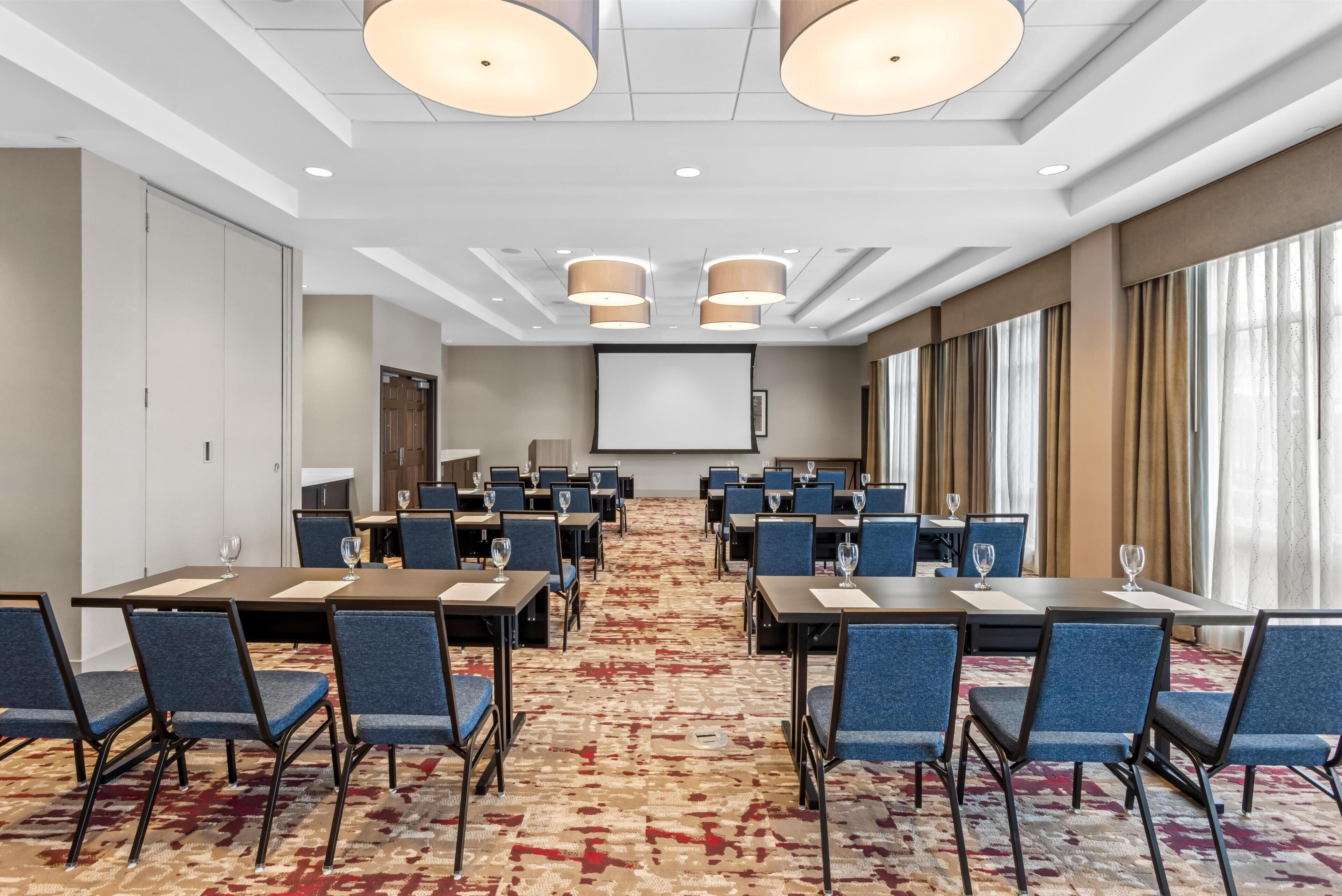 Une salle de conférence équipée de plusieurs tables et chaises, d’un écran de projection et d’un tapis à motifs.
