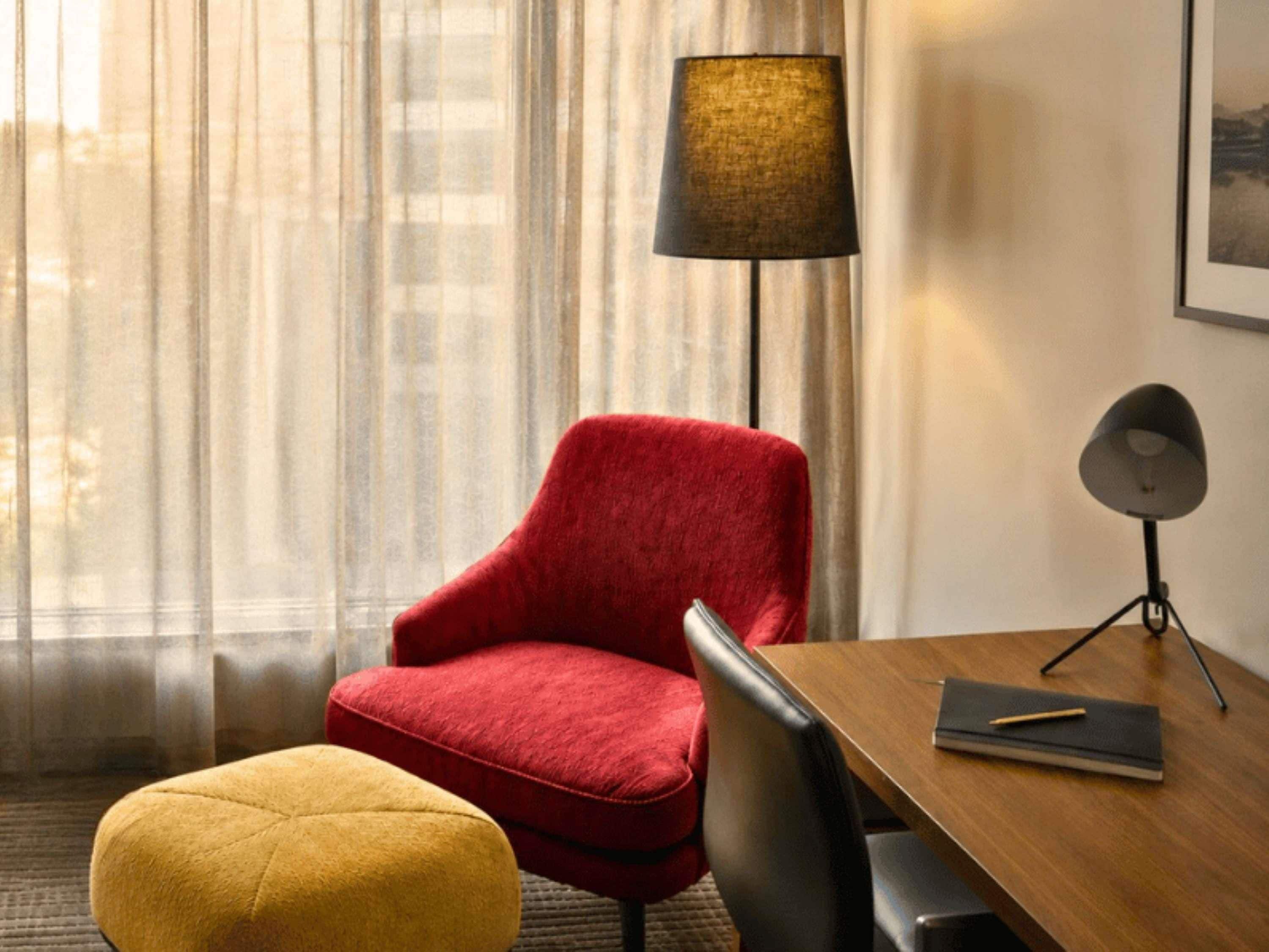 A hotel room with a wooden desk, a red chair, a yellow ottoman, a black lamp, and sheer curtains.