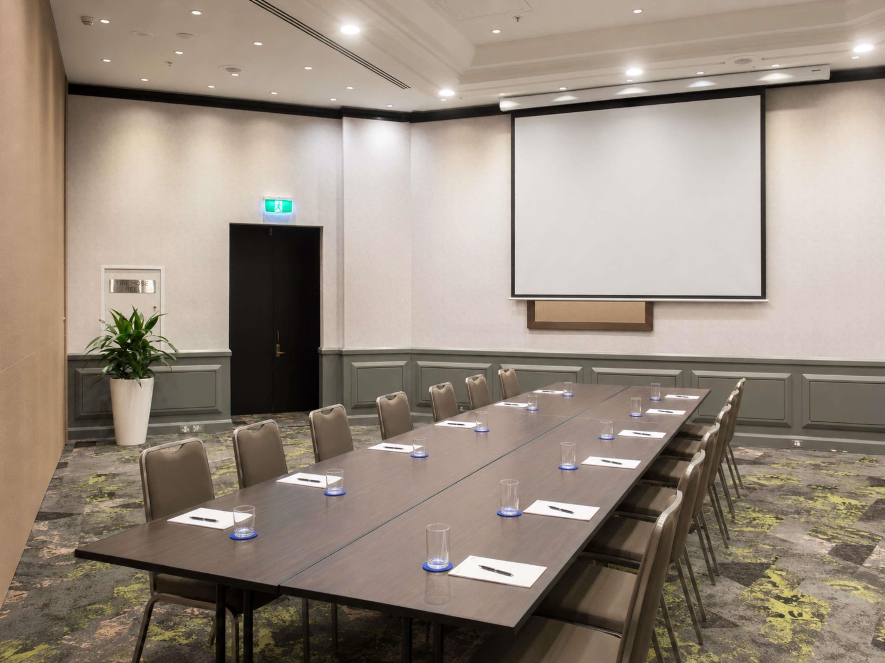 A conference room with a long table, chairs, a projector screen, and a door marked 'Fire Hose Reel.'