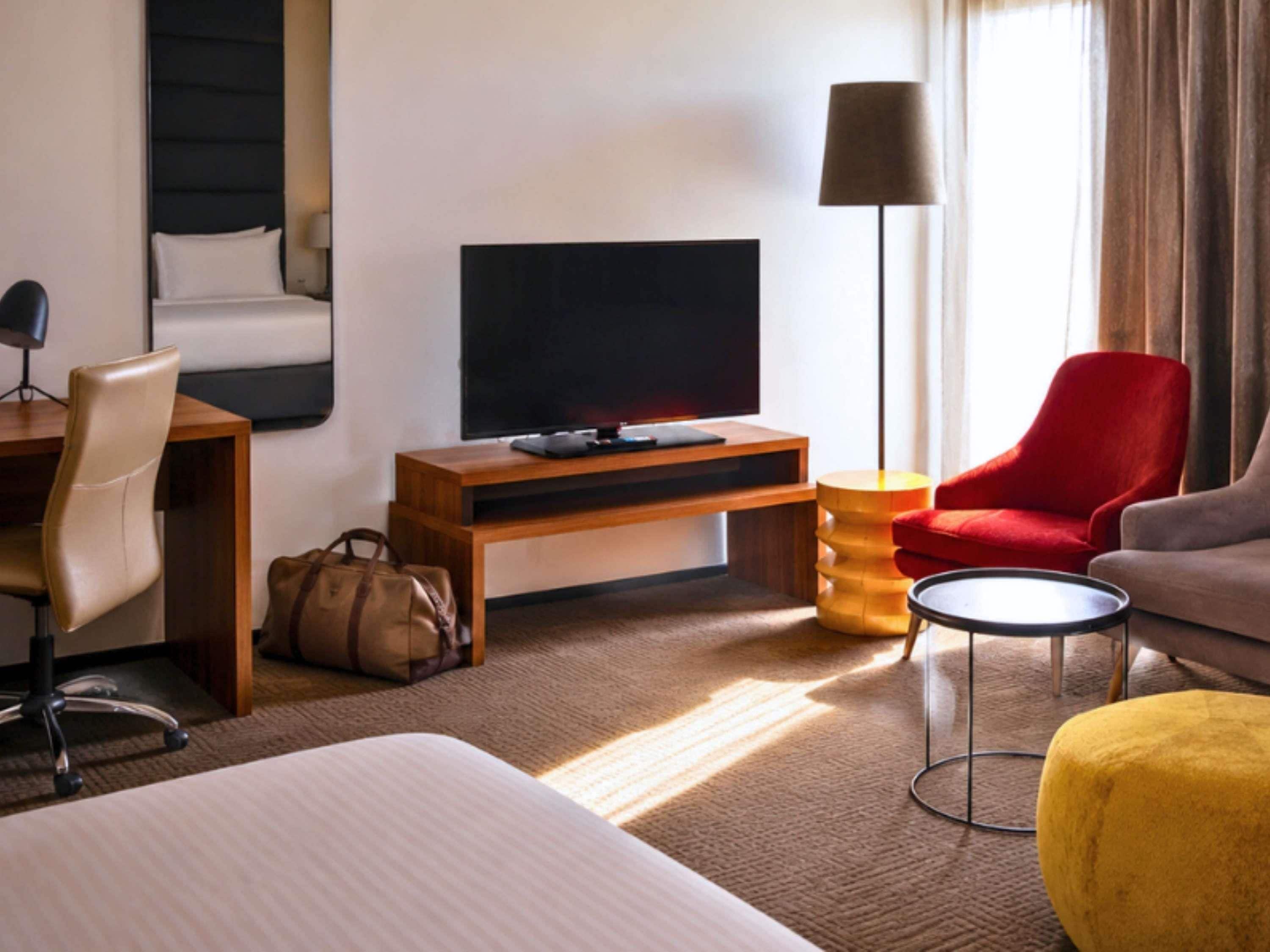 A hotel room with a bed, a desk, a TV, a red chair, a yellow ottoman, and a lamp.