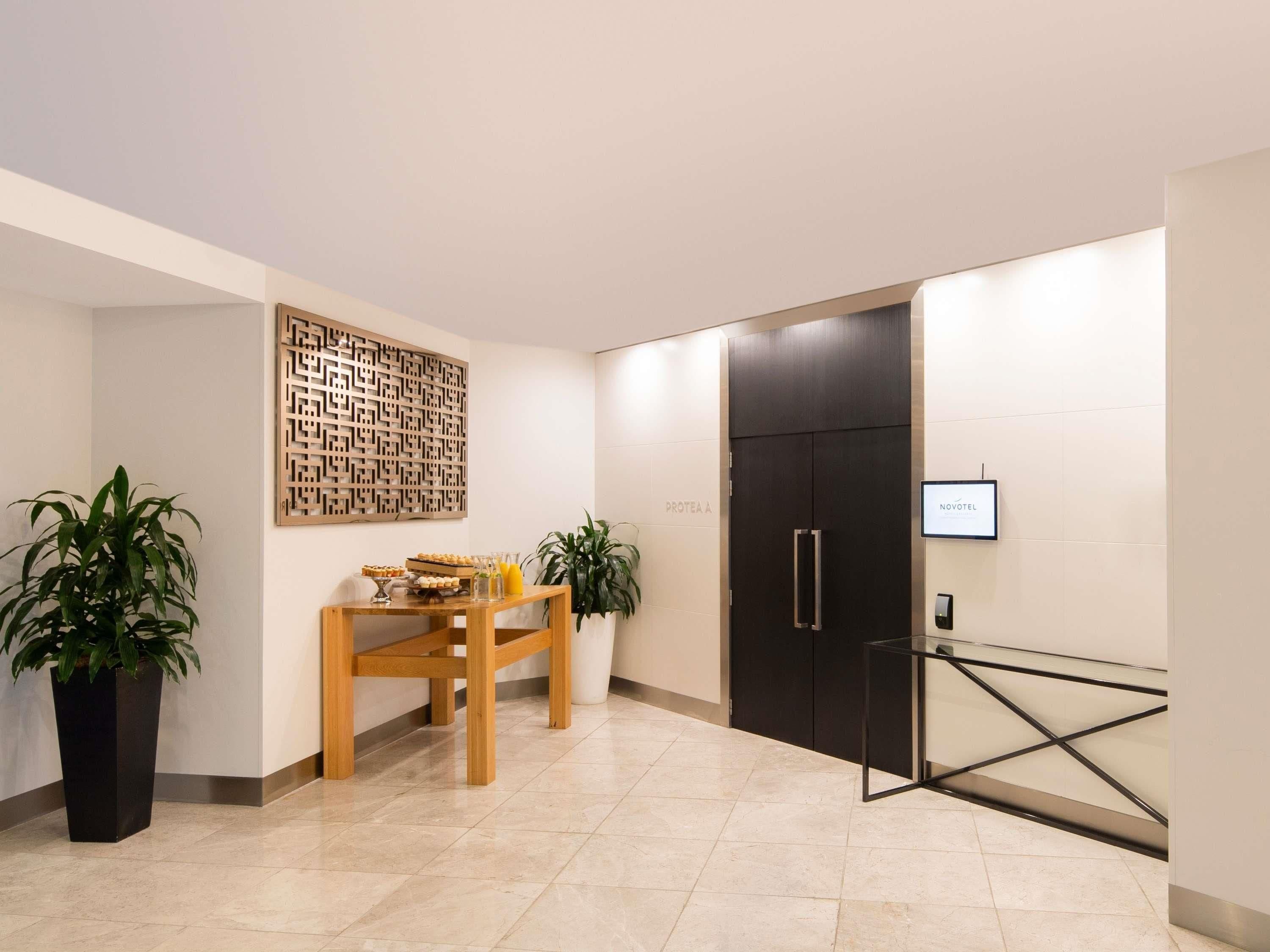A hotel lobby with a reception desk, a wall-mounted TV, a black door, and potted plants.
