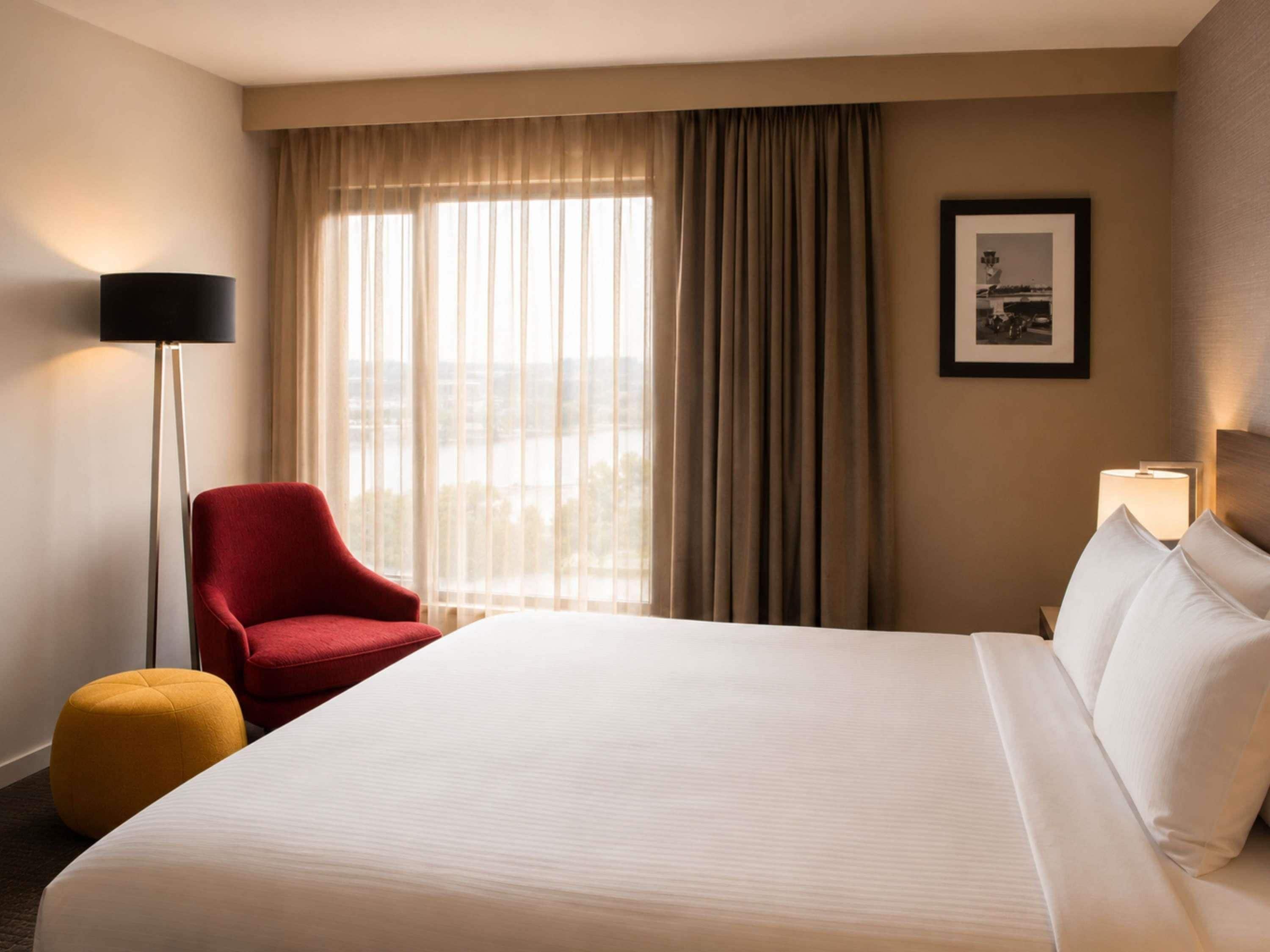 A hotel room with a bed, a red armchair, a yellow ottoman, a floor lamp, a window with curtains, and a framed picture on the wall.