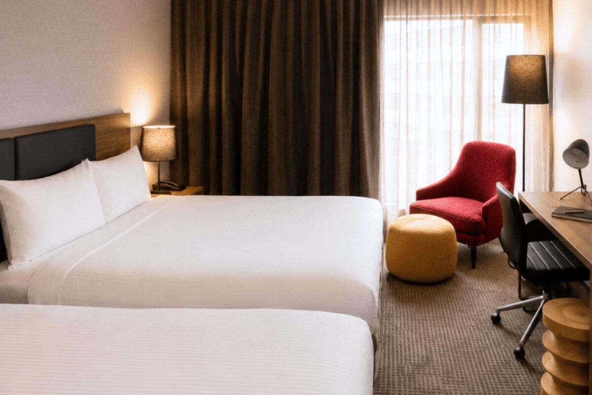 A hotel room with two beds, a desk, a chair, a lamp, and a window with curtains.