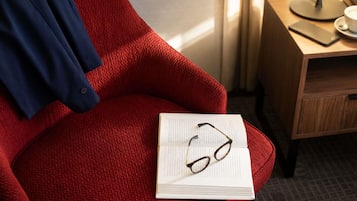 Una silla roja con textura, con un libro y gafas encima, un teléfono inteligente sobre una mesa y una lámpara al fondo.