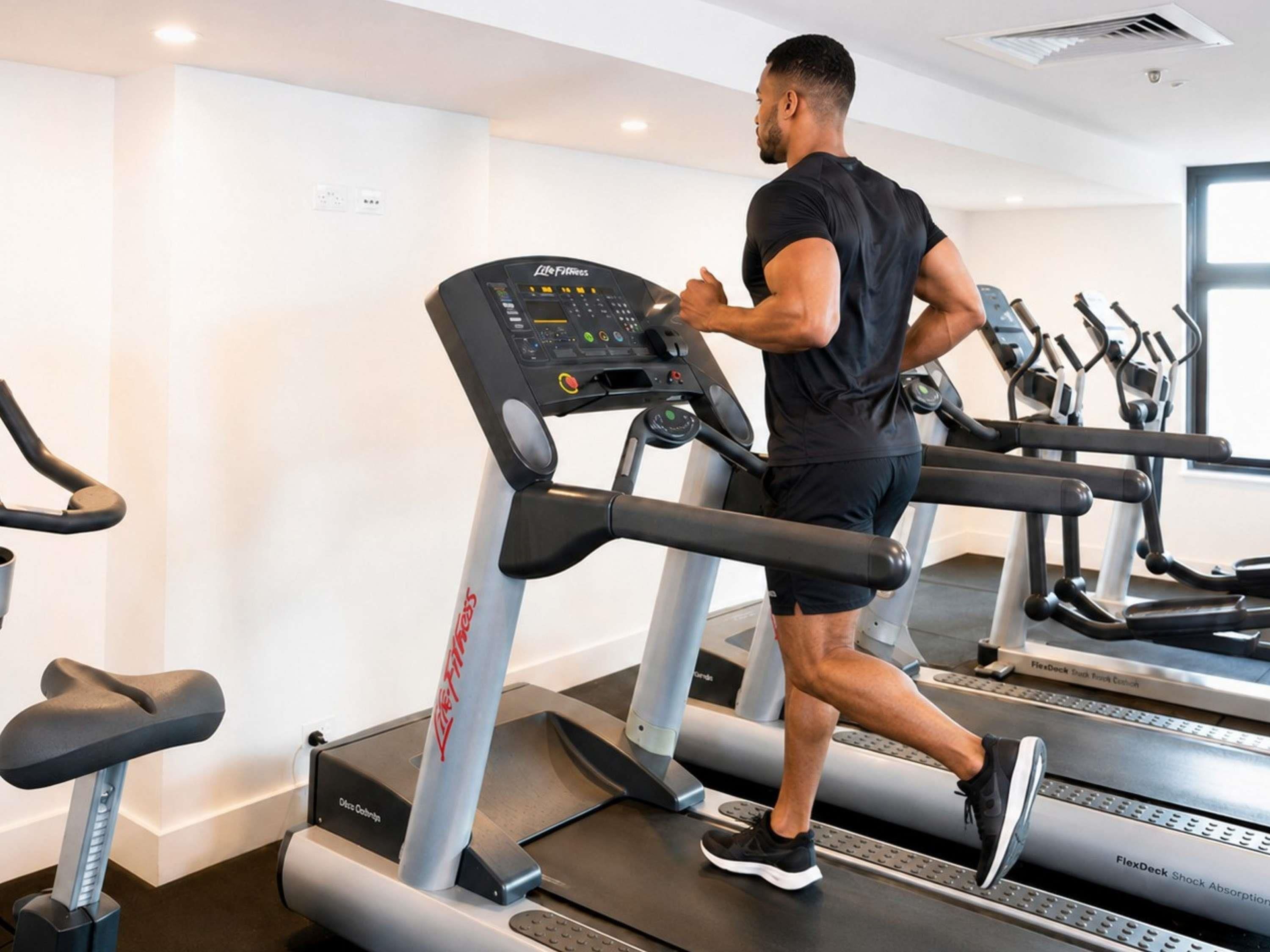Un homme court sur un tapis de course dans une salle de sport équipée de plusieurs tapis de course et d’appareils d’exercice.
