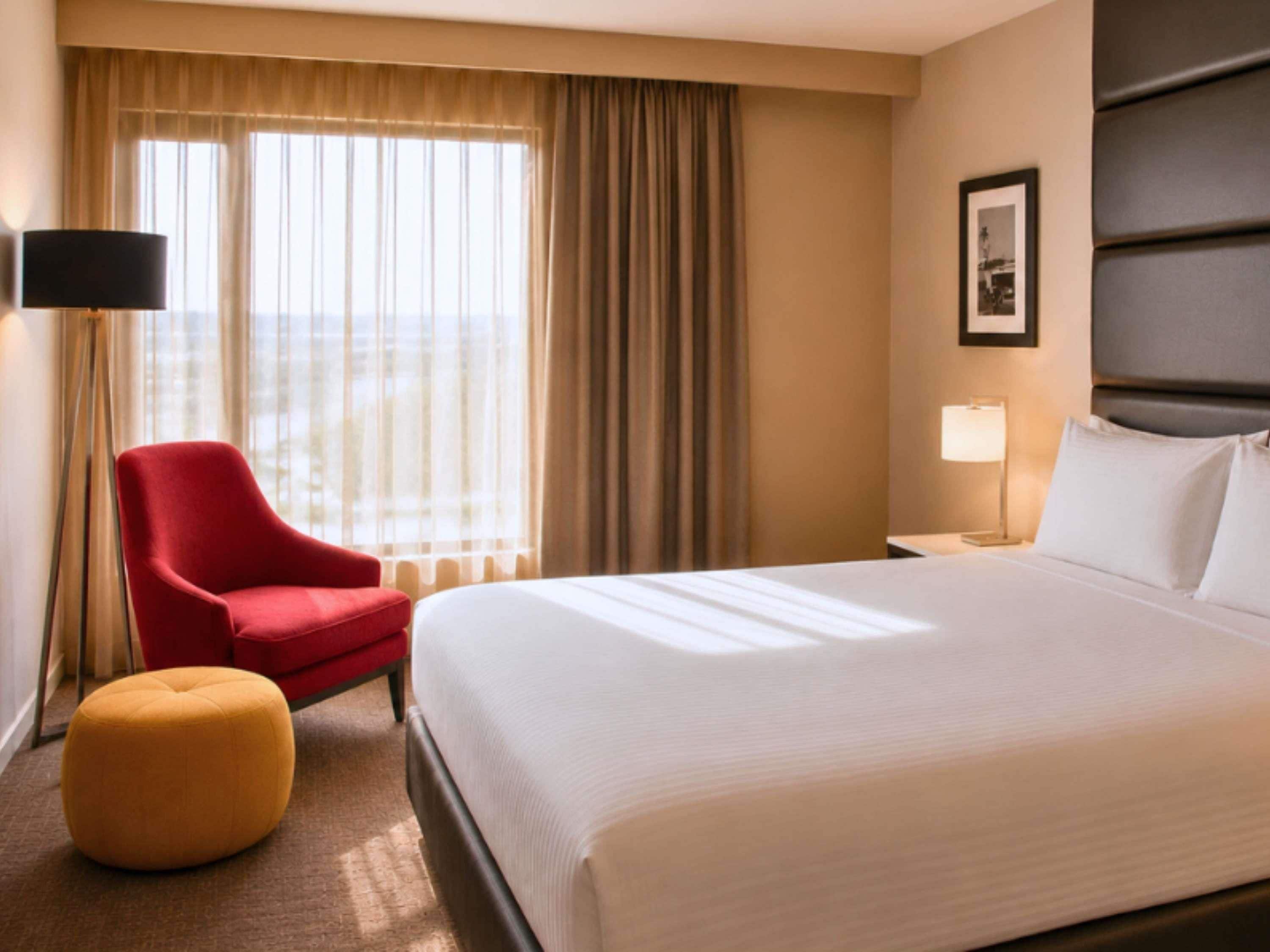 Une chambre d’hôtel avec un lit, un fauteuil rouge, un pouf jaune, une table de chevet avec une lampe et une fenêtre avec des rideaux.