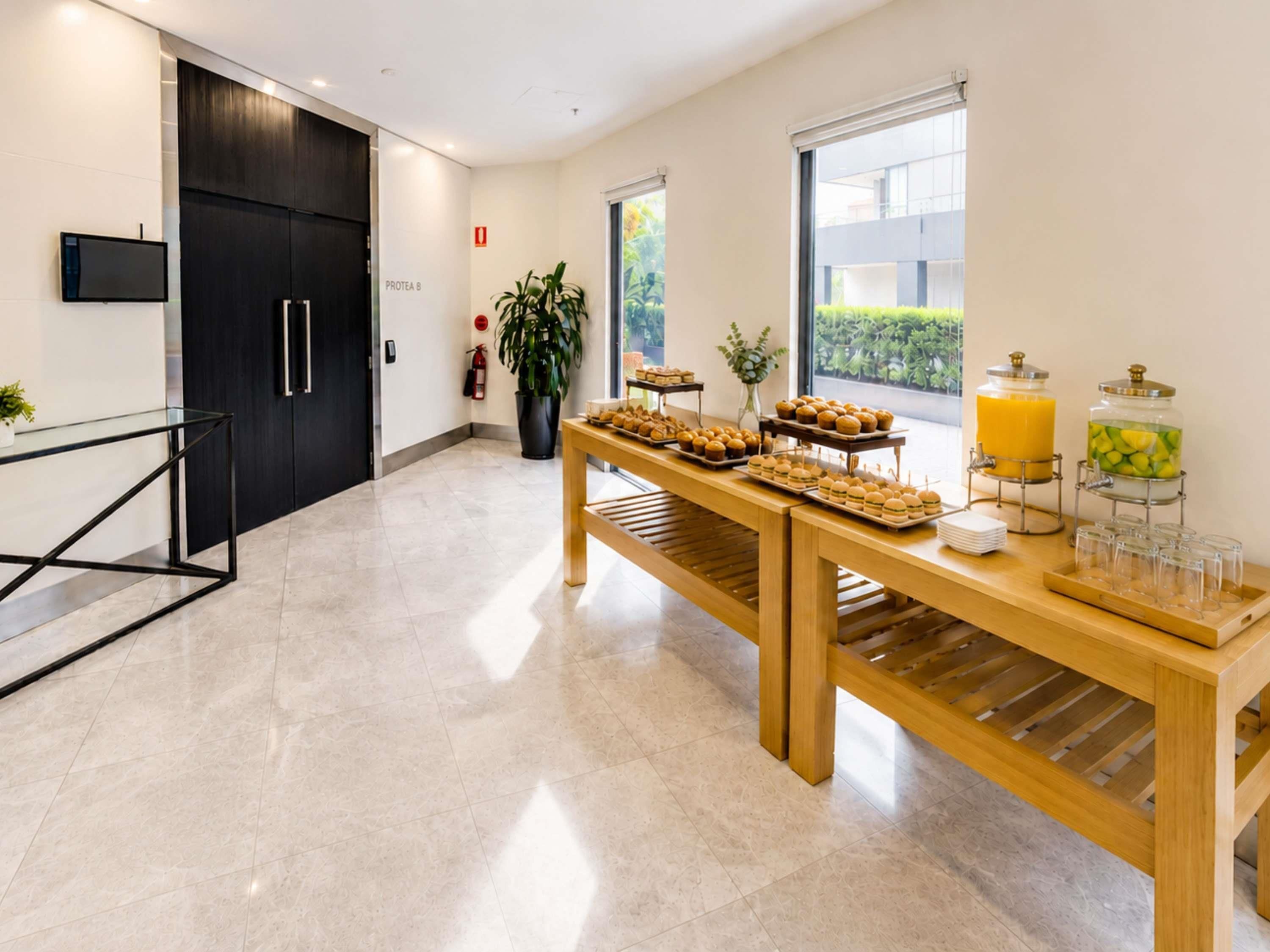 A modern hotel lobby with a reception desk, a buffet table with food and drinks, and a large window offering a view of the outdoors.