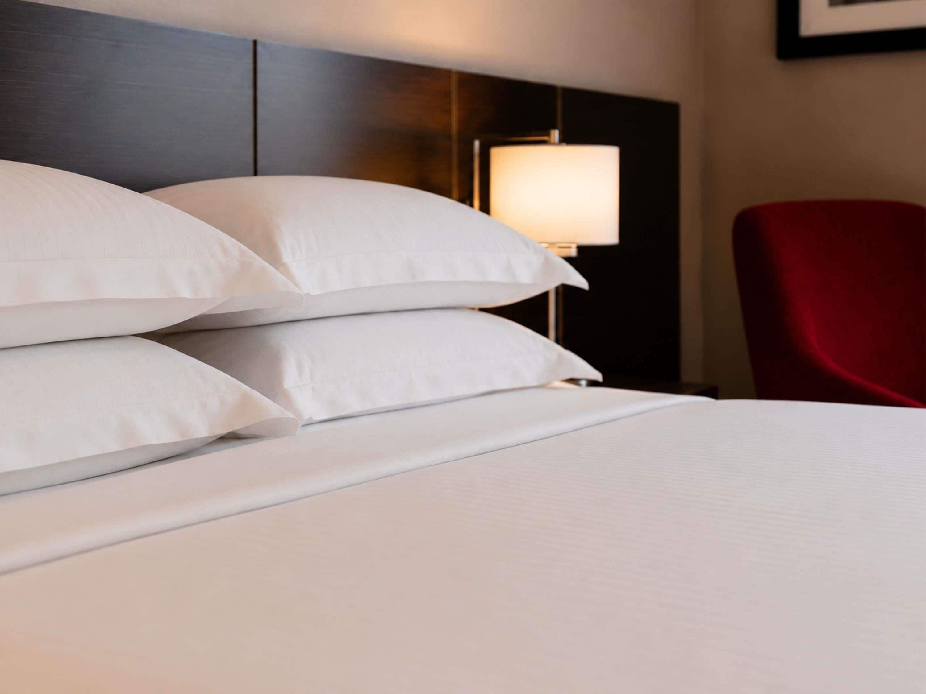 Un lit bien fait, avec du linge de lit blanc et des oreillers, une chaise rouge et une lampe fixée au mur.