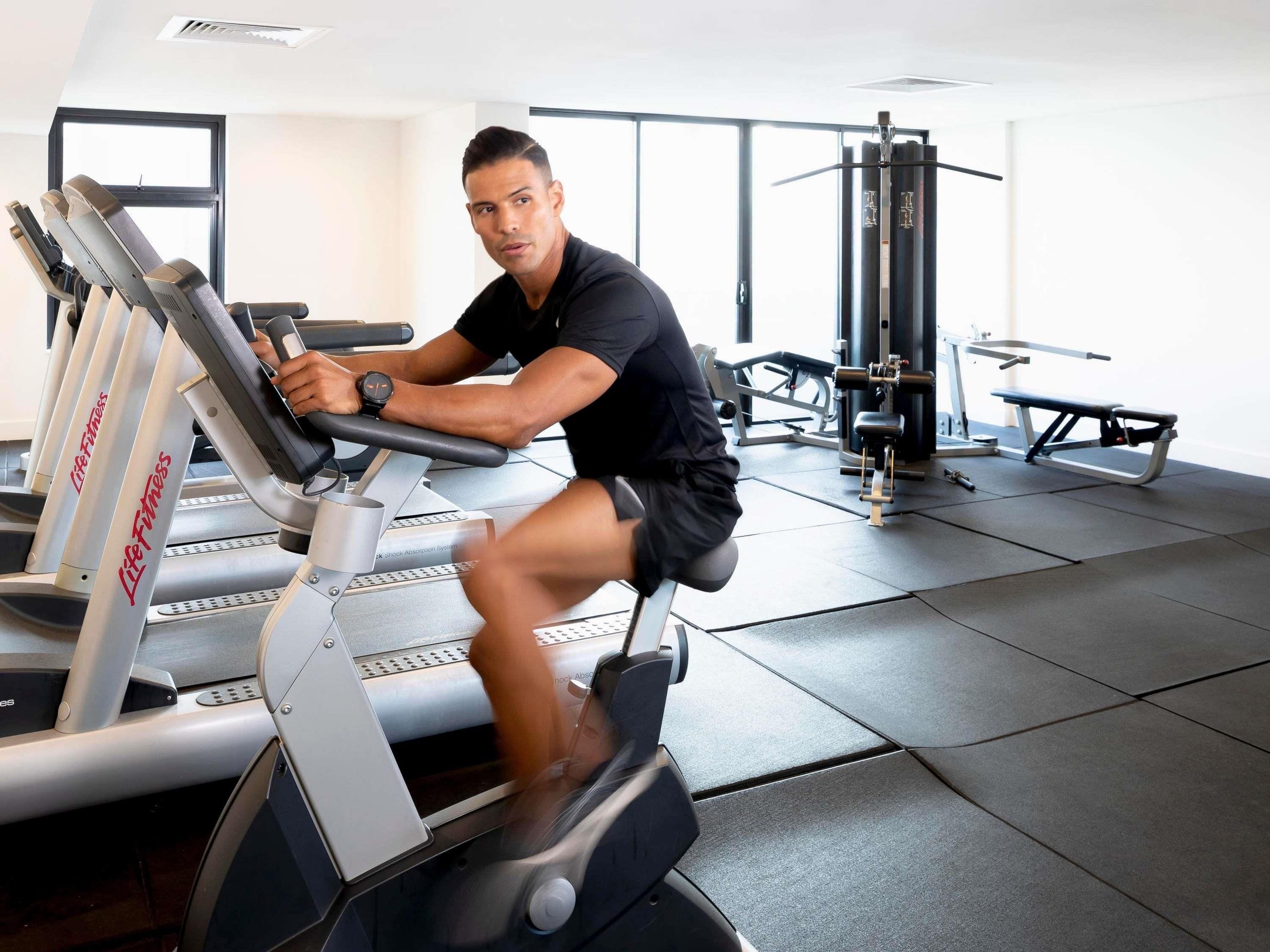 Una persona usando una máquina elíptica en un gimnasio, con cintas de correr y una banca de pesas al fondo.