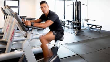 Una persona usando una máquina elíptica en un gimnasio, con cintas de correr y una banca de pesas al fondo.