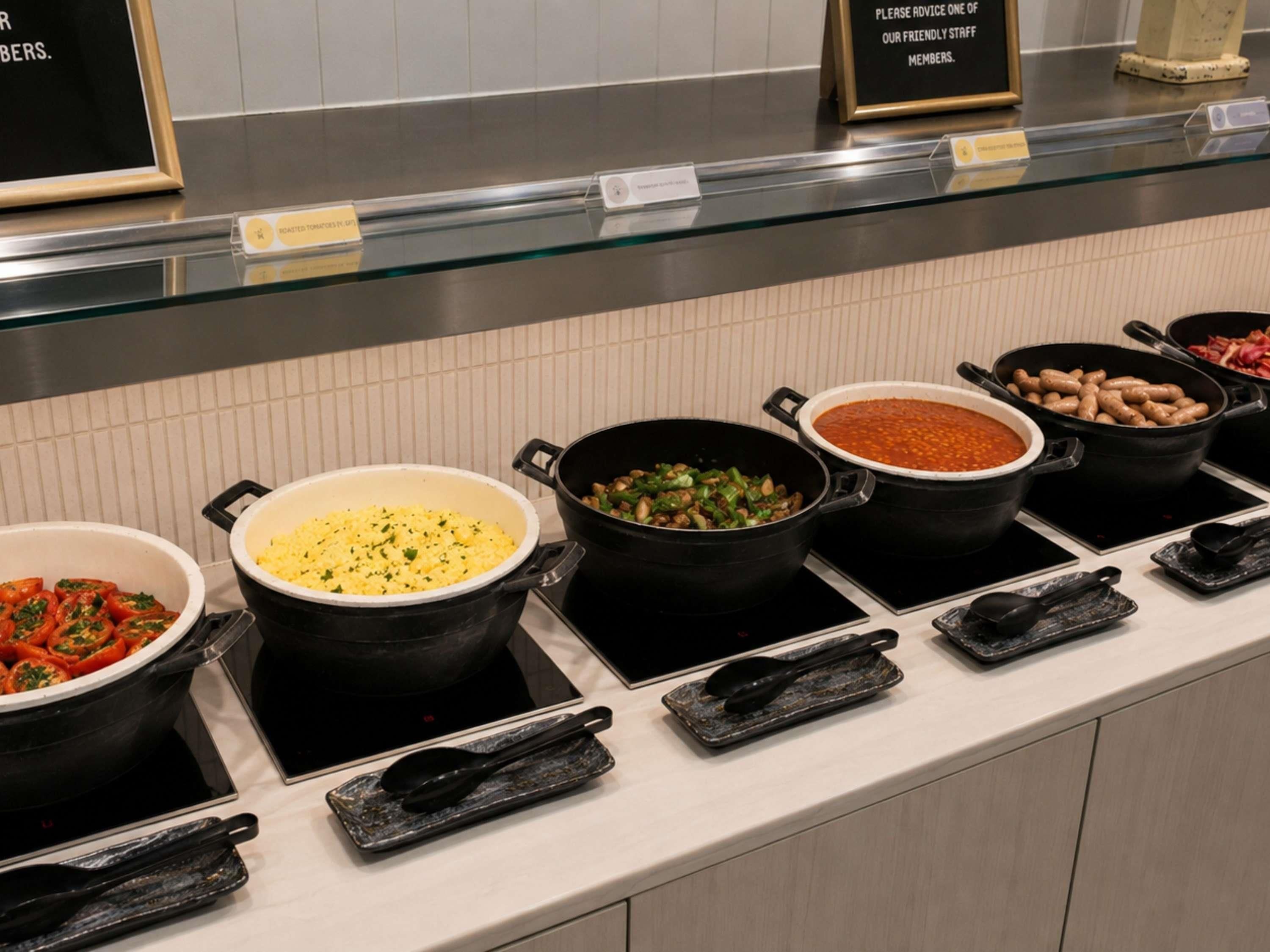 A buffet counter with various dishes in black pots, a sign advising to seek staff advice, and a tiled wall in the background.