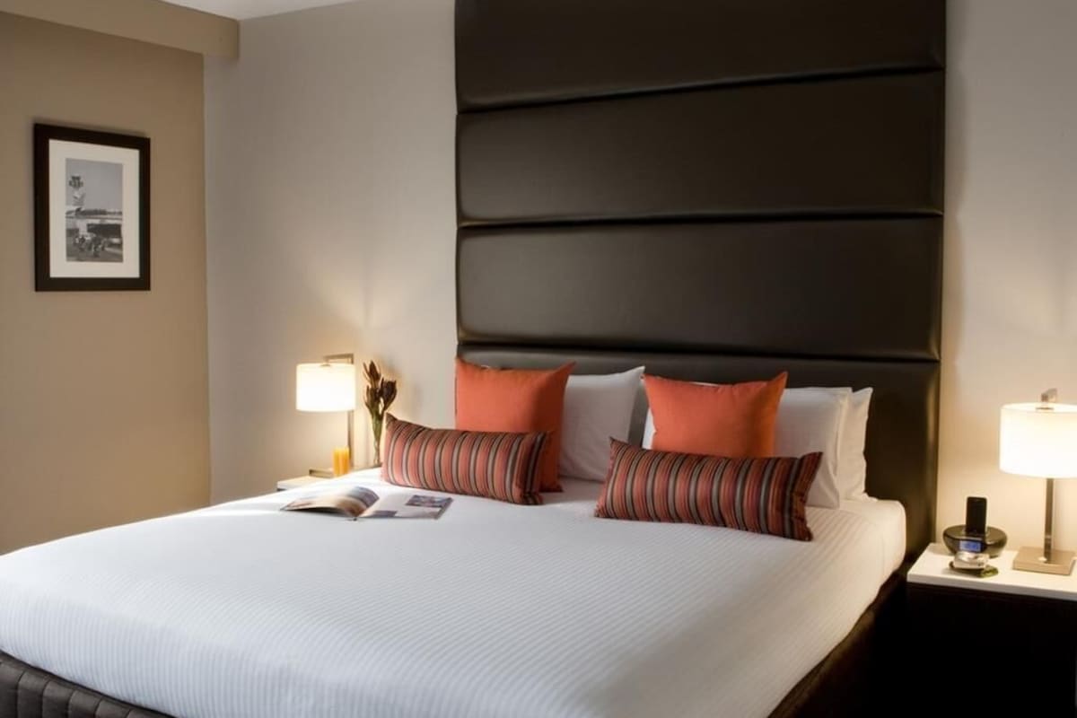 A hotel room with a large bed, two bedside lamps, a framed picture on the wall, and a dark headboard.