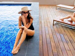 A person sitting by a pool with a wooden deck and a lounge chair.
