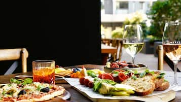 A dining table with a pizza, a glass of red drink, a cheese board with various cheeses and olives, and two glasses of white wine.