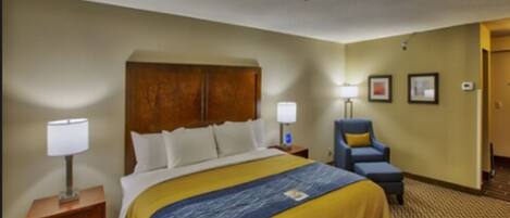 A hotel room with a large bed, a nightstand with a lamp, a chair, and a wall-mounted TV.