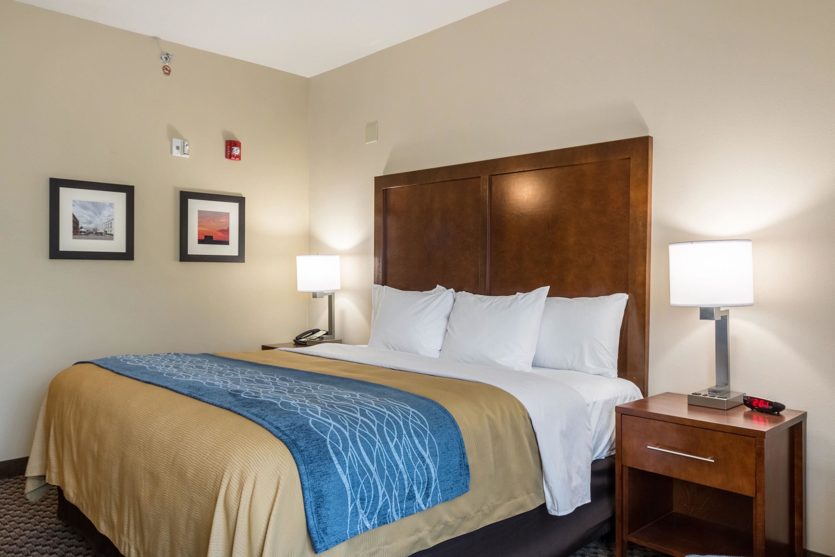 Una habitación de hotel con una cama grande, dos lámparas de noche, una mesita de noche con un reloj y dos cuadros en la pared.