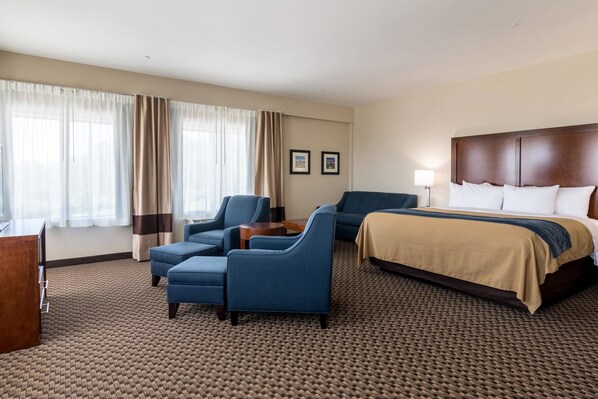 A hotel room with a large bed, two blue armchairs, a small table, and a desk with a lamp.