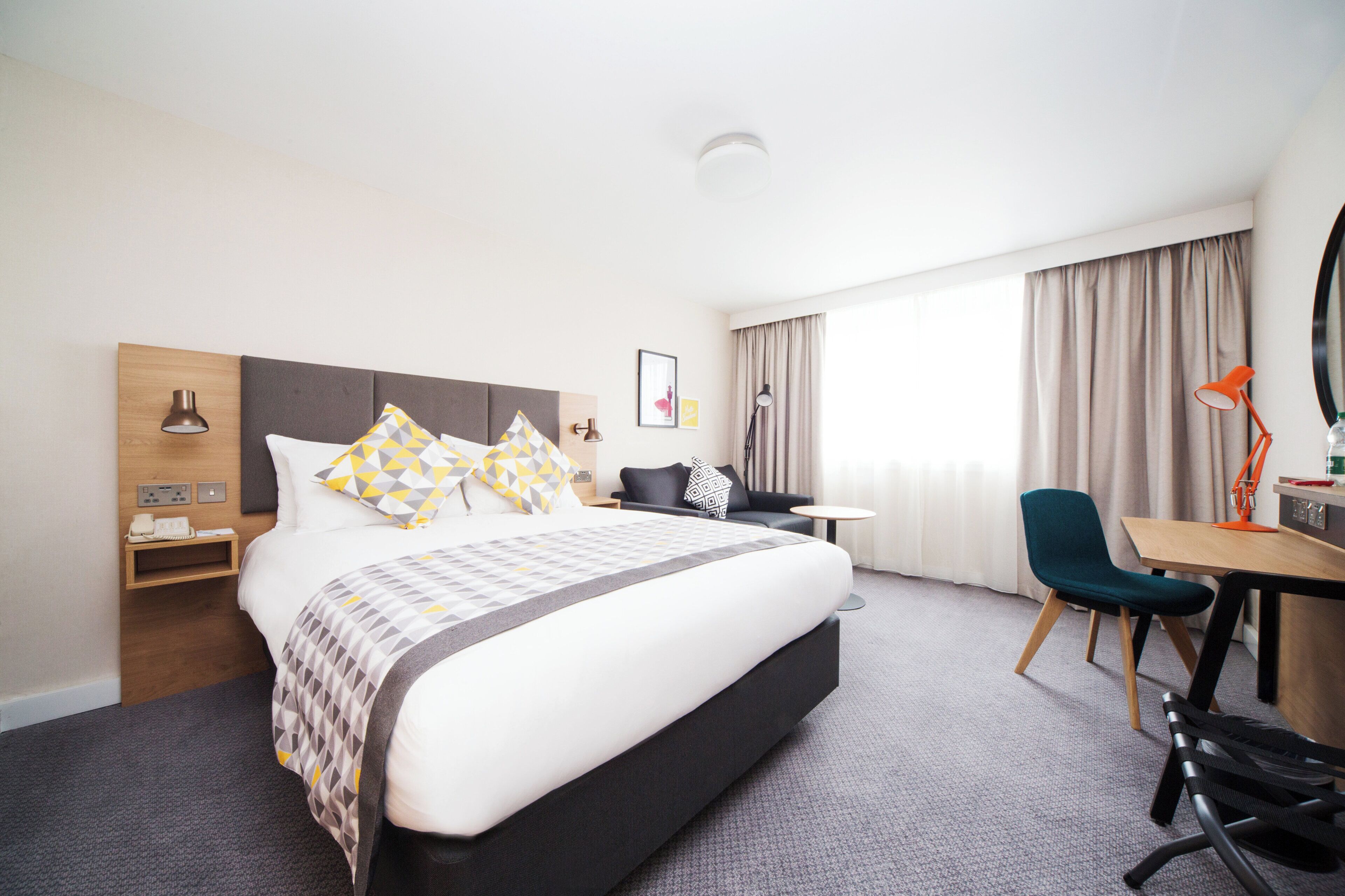 A hotel room with a large bed, a desk, a chair, a small table, and a window with curtains.