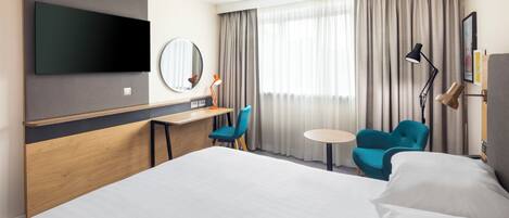 Ein Hotelzimmer mit einem großen Bett, einem Schreibtisch mit Stuhl, einem kleinen runden Tisch und einem an der Wand befestigten Fernseher.