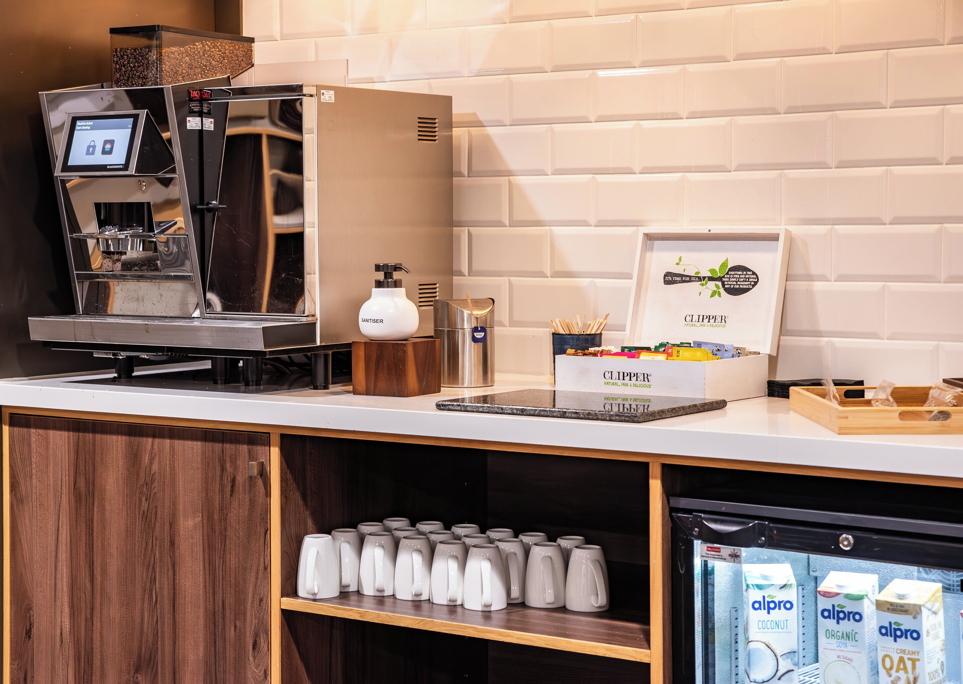 A coffee station with an espresso machine, a milk frother, and a Clipper brand coffee grinder. There are also milk cartons and a refrigerated display of Alpro products.