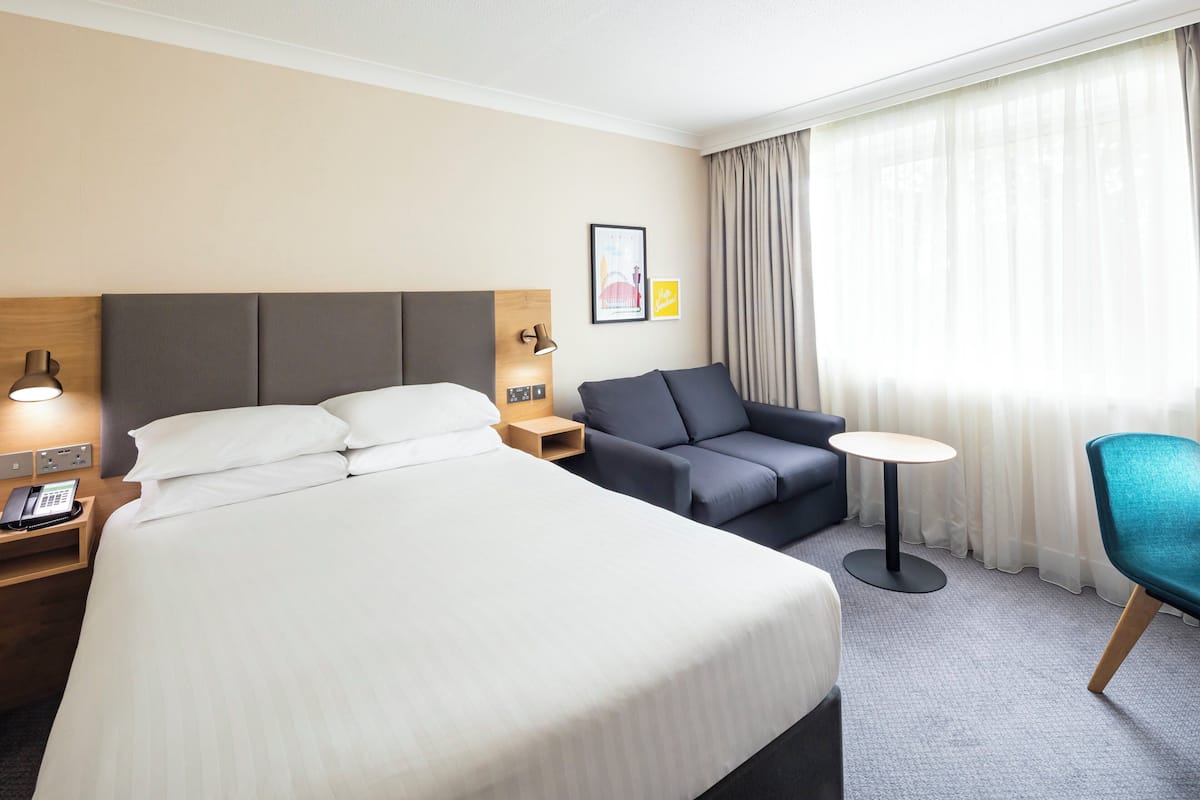 A hotel room with a large bed, a sofa, a small table, and a chair. There is a nightstand with a lamp and a phone.