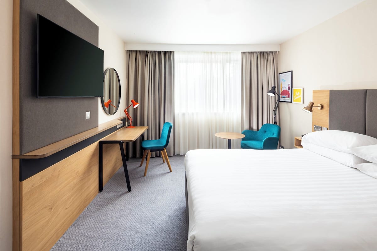 A hotel room with a bed, a desk with a chair, a TV mounted on the wall, a small table, and a window with curtains.