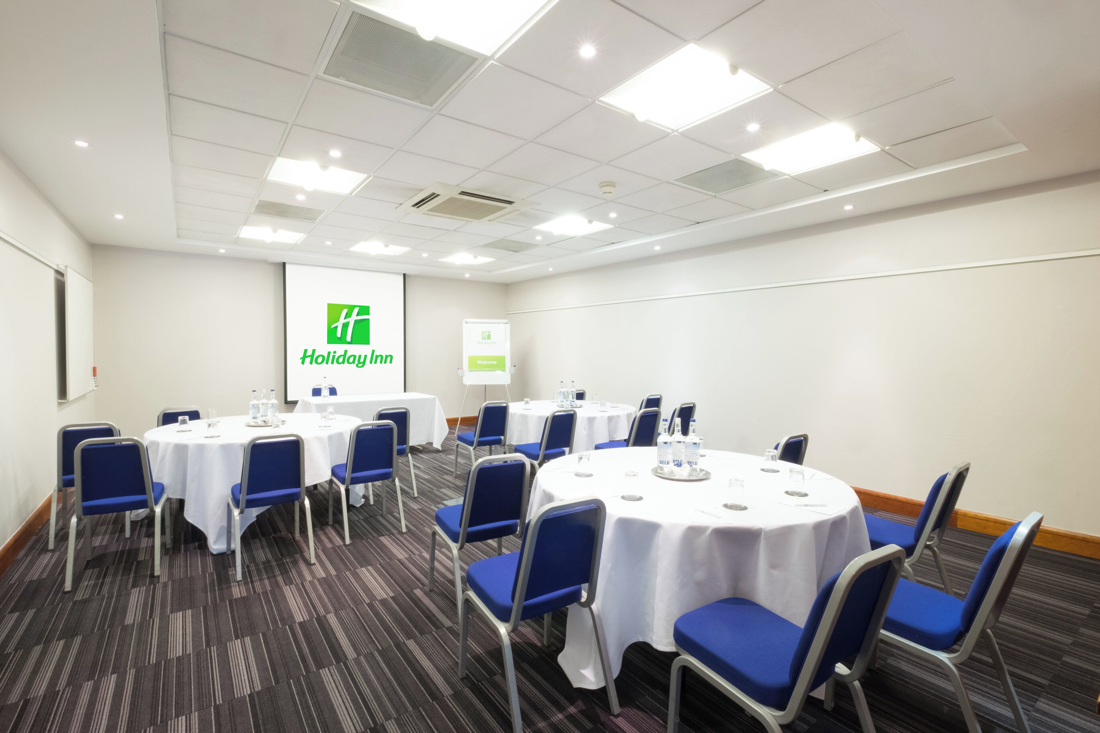 A conference room with round tables, blue chairs, and a large screen displaying the Holiday Inn logo.