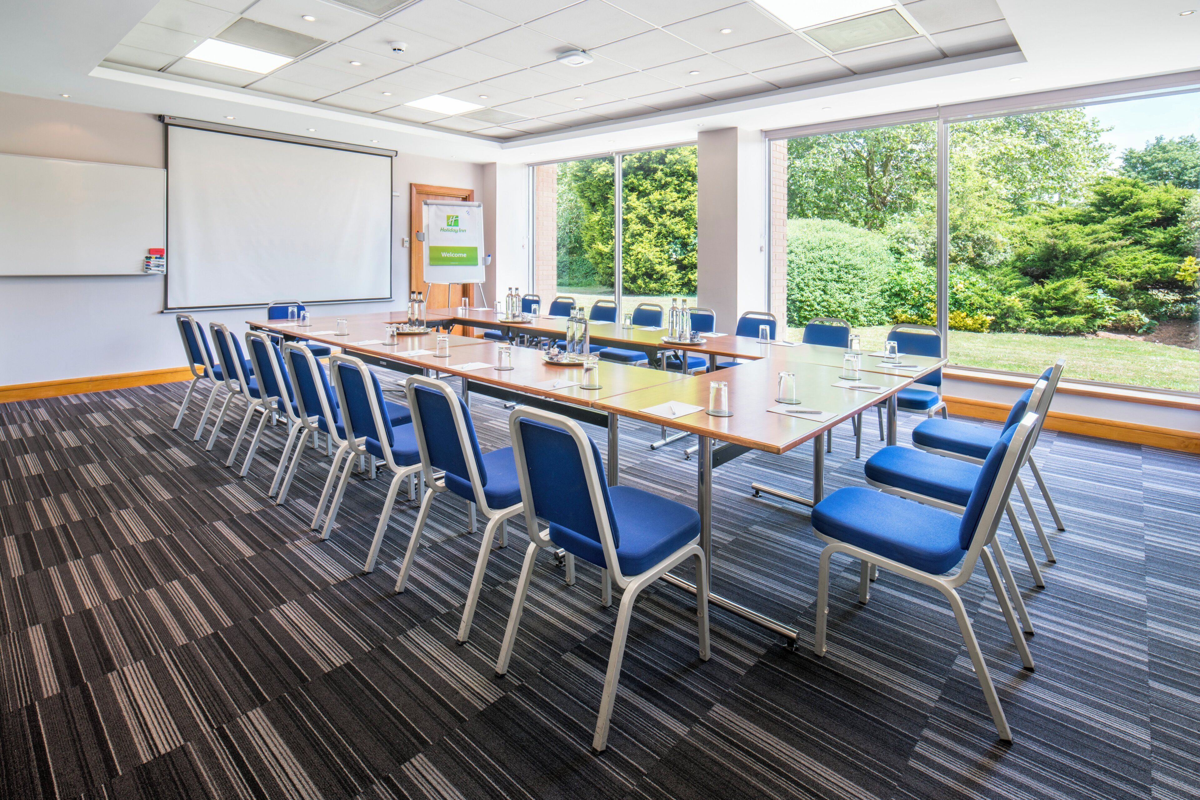 A conference room with a long table, blue chairs, a projector screen, and a view of greenery outside.