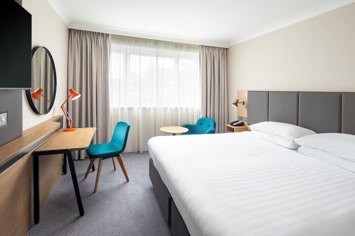 A hotel room with a bed, a desk, two chairs, a small table, a TV, and a window with curtains.