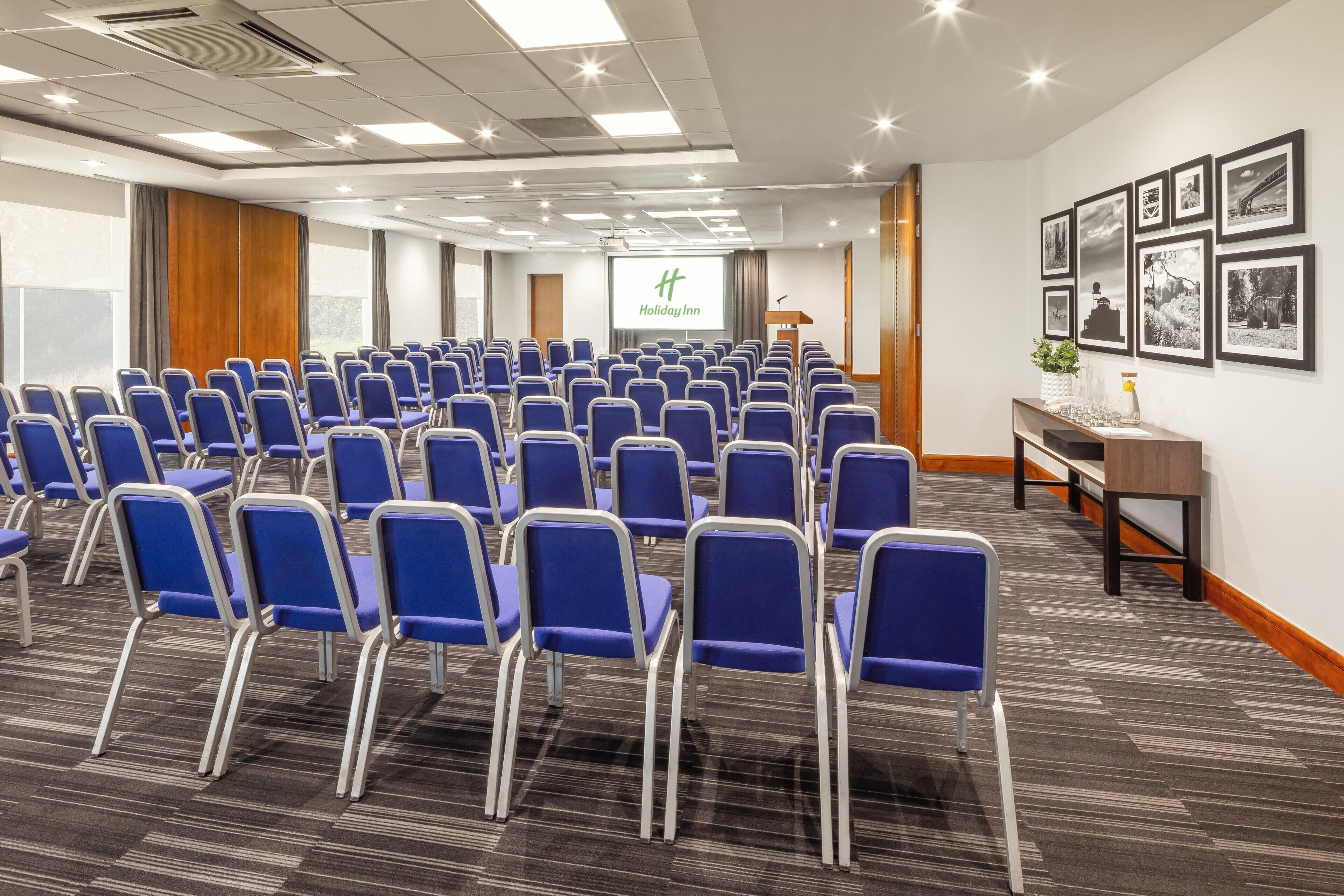 Een vergaderzaal met rijen blauwe stoelen, een spreekgestalte, een scherm waarop 'Holiday Inn' staat en ingelijste foto's aan de muur.