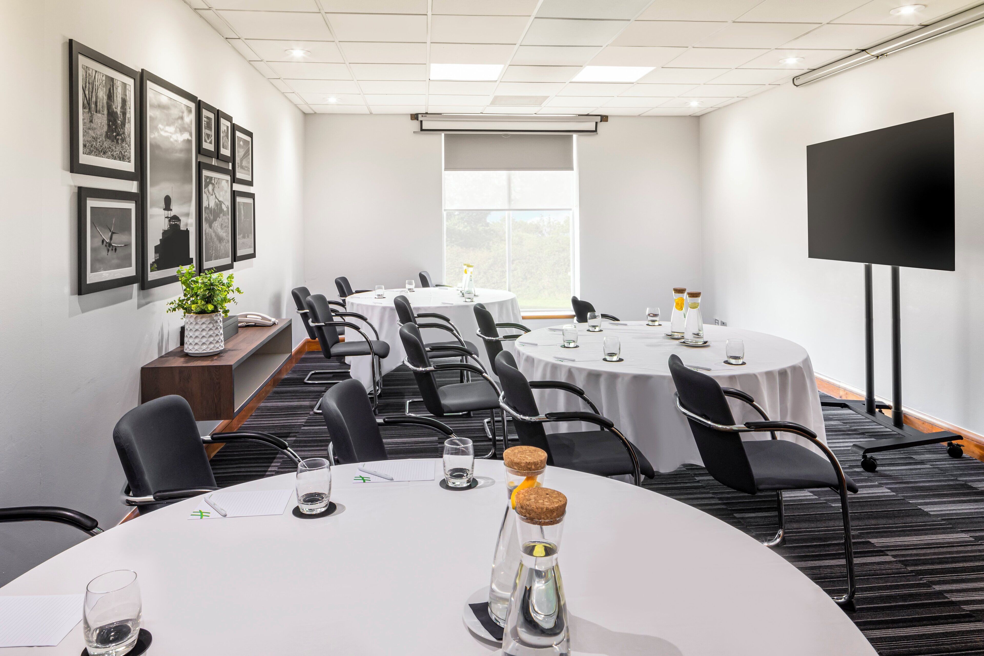 A conference room with round tables, black chairs, a projector screen, and framed pictures on the wall.