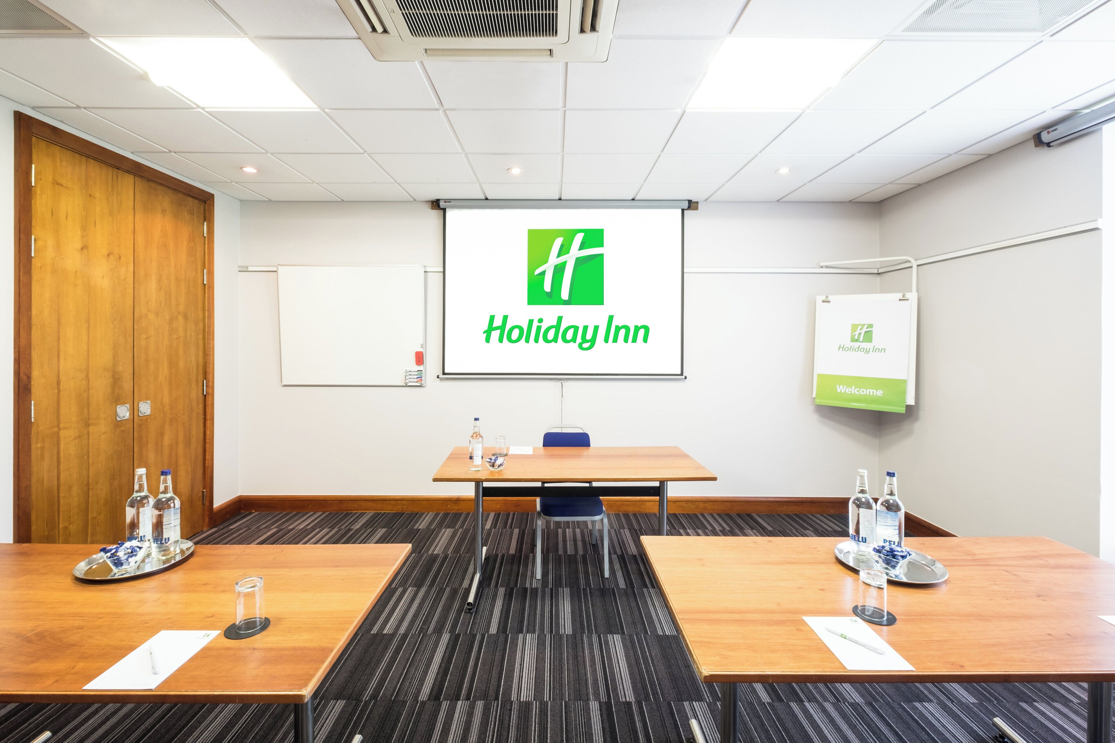 A conference room with a Holiday Inn logo on the screen, a whiteboard, and a welcome sign.
