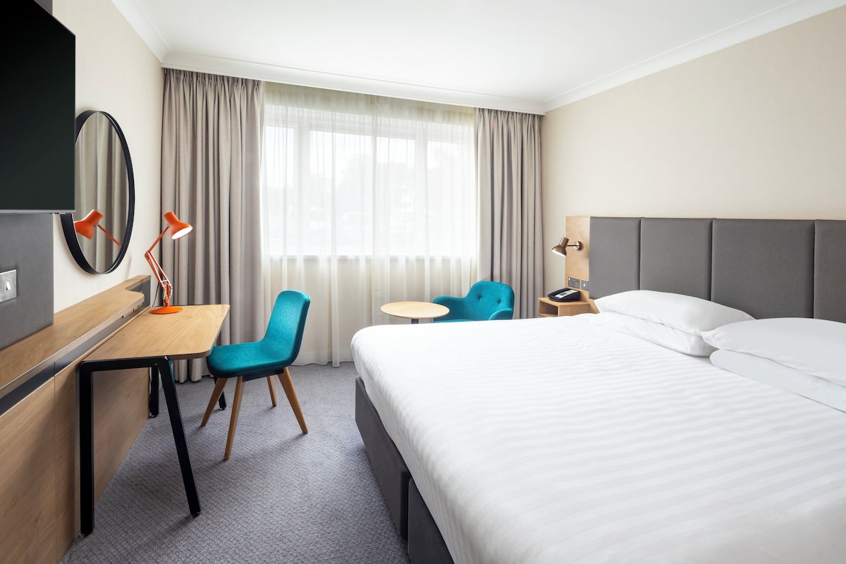 A hotel room with a bed, a desk, two chairs, a small table, a TV, and a window with curtains.