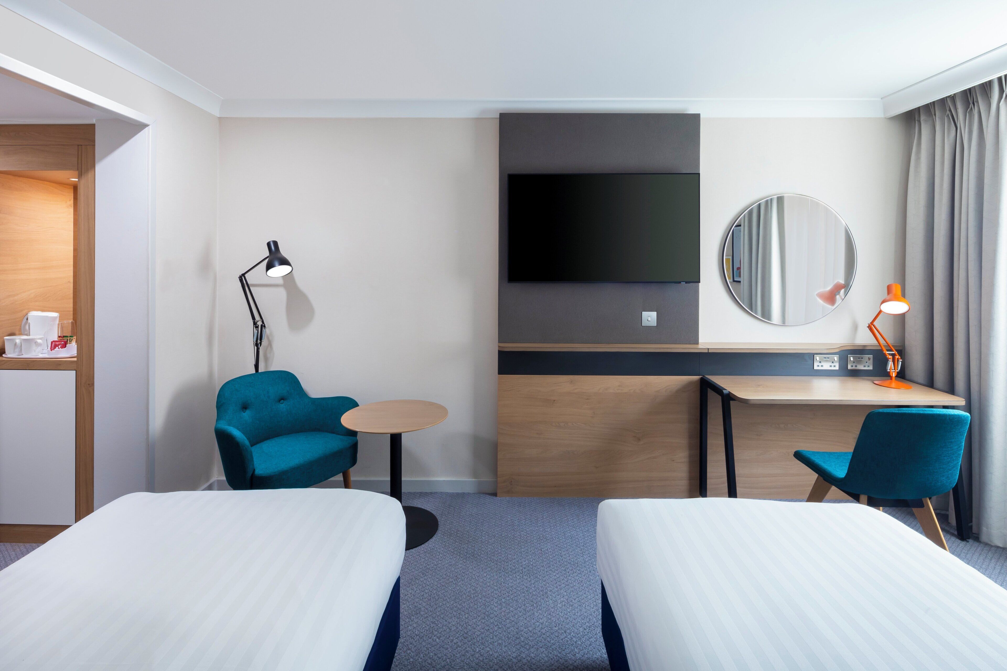 A hotel room with two single beds, a desk with a chair, a TV, a round mirror, and a small table with a lamp.