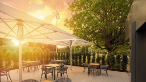 An outdoor patio with tables and chairs under a large umbrella, surrounded by greenery and a flag. - Holiday Inn London-Gatwick Airport by IHG (Horley)