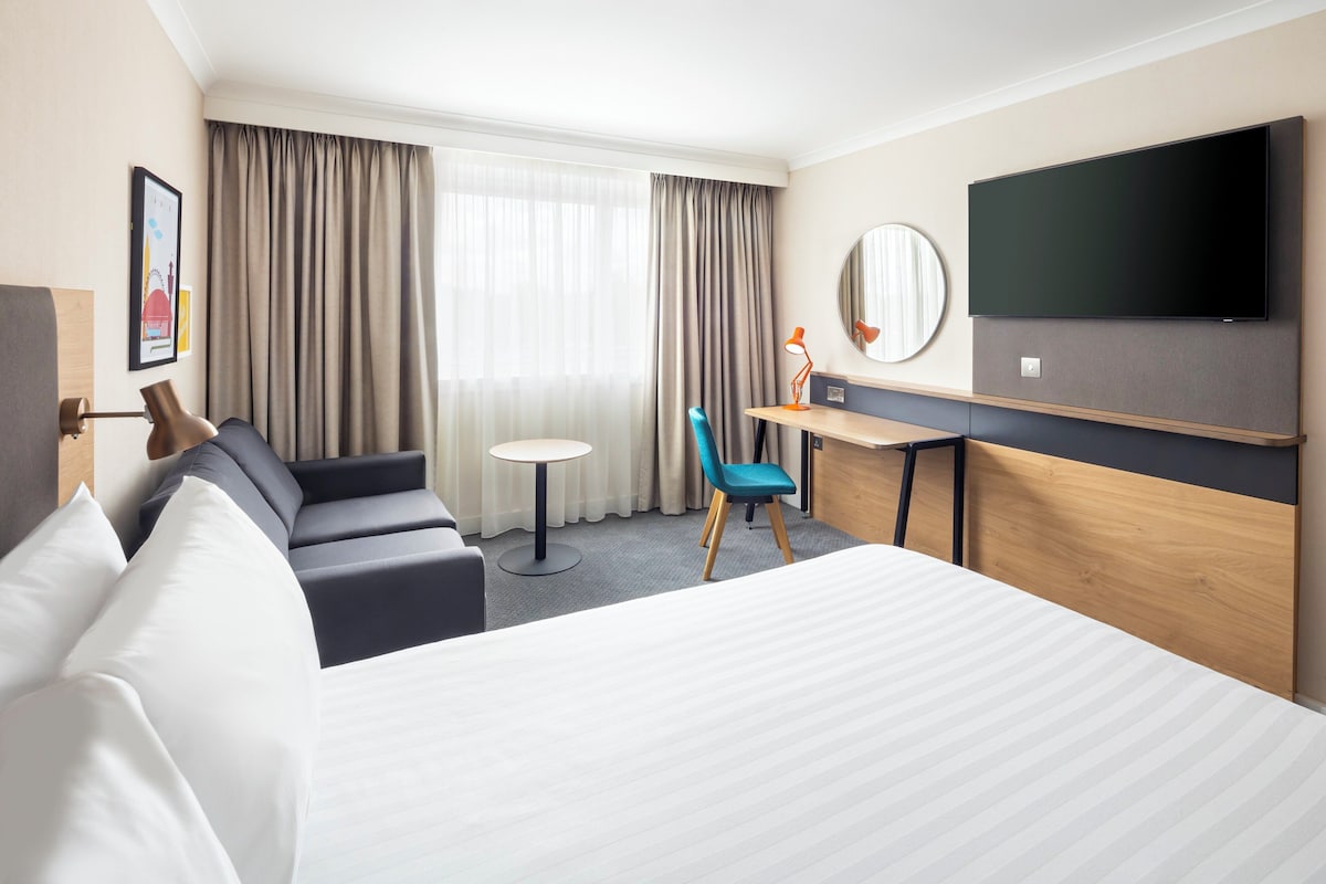 A hotel room with a large bed, a desk, a chair, a TV, and a window with curtains.