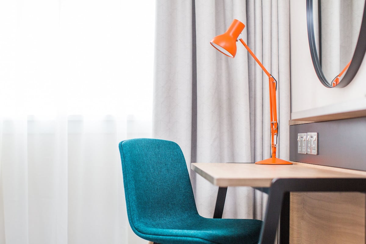 A teal chair, a wooden desk, an orange desk lamp, and a wall-mounted mirror.
