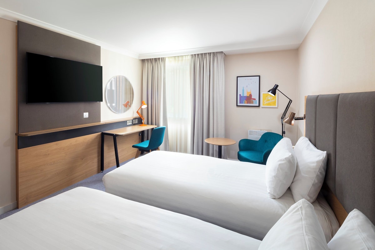A hotel room with two beds, a desk, a chair, a TV, a lamp, and a small table.