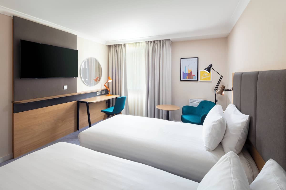 A hotel room with two beds, a desk, a chair, a TV, a lamp, and a small table.