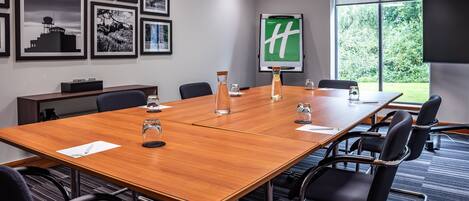 A conference room with a long wooden table, black chairs, framed pictures on the wall, a projector screen with a green logo, and a large window with a view of trees.